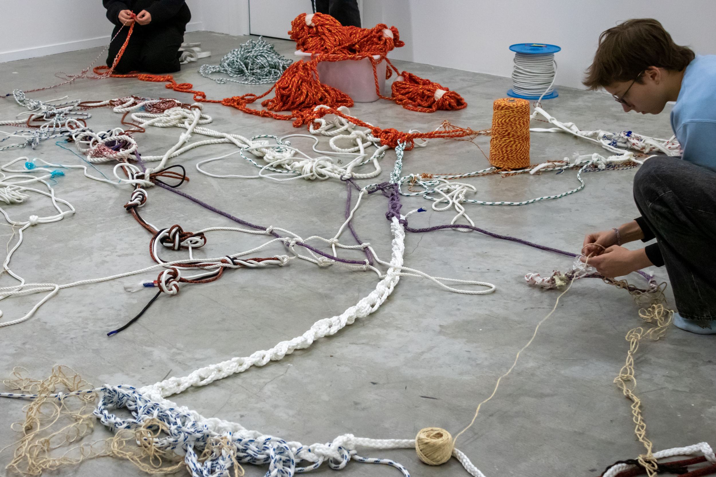 A large mesh of ropes is laying on the floor. At the edges of the mesh, two people are seen sitting tying ropes together.