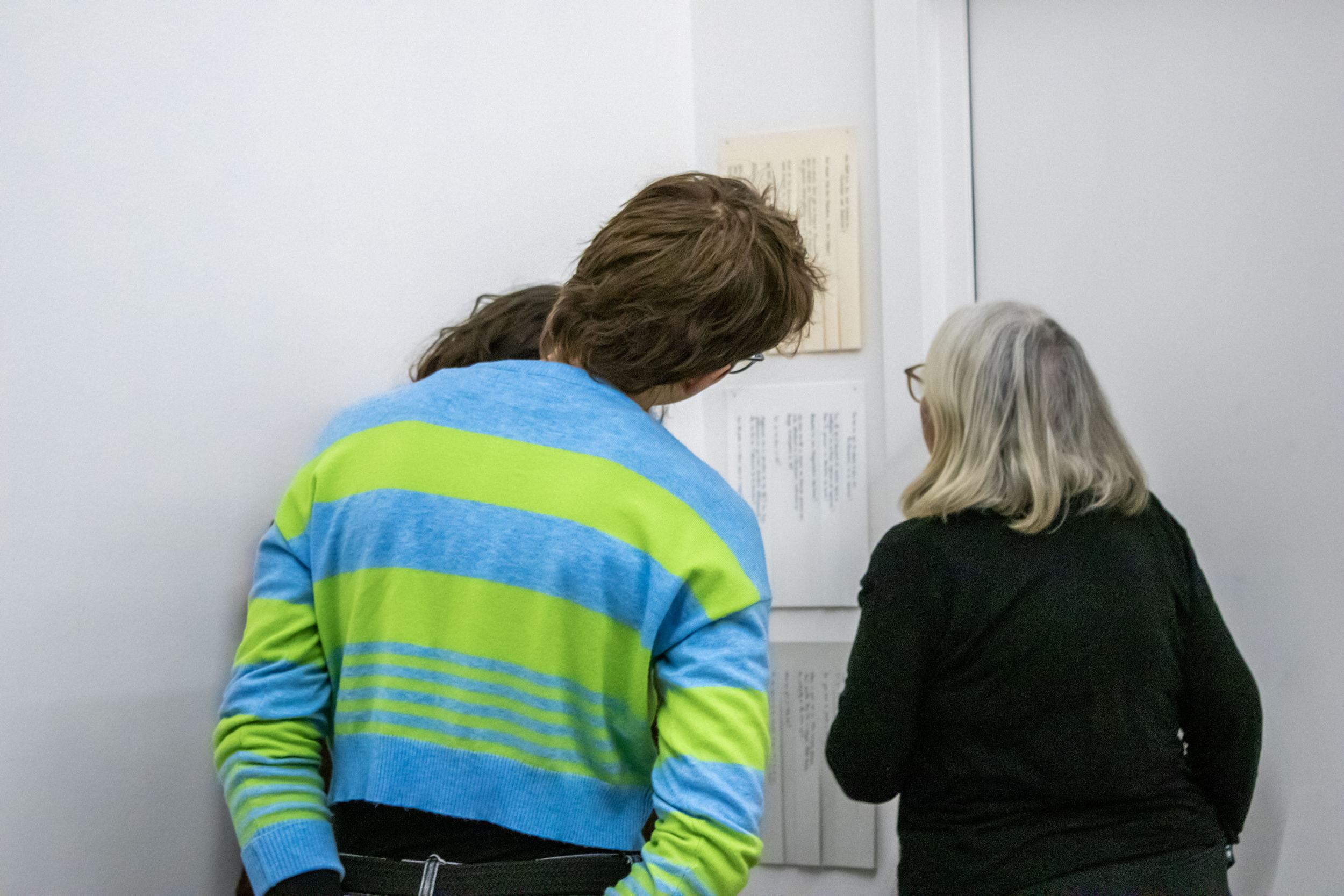 Three people are standing looking at three blocks of papers nailed to the wall one under another. The papers are cut to form vertical stripes that can be torn out, each stripe is printed with a different question. Each block uses a different language and has a slightly different paper color: the top block is beige, the middle one white, the bottom one light grey.