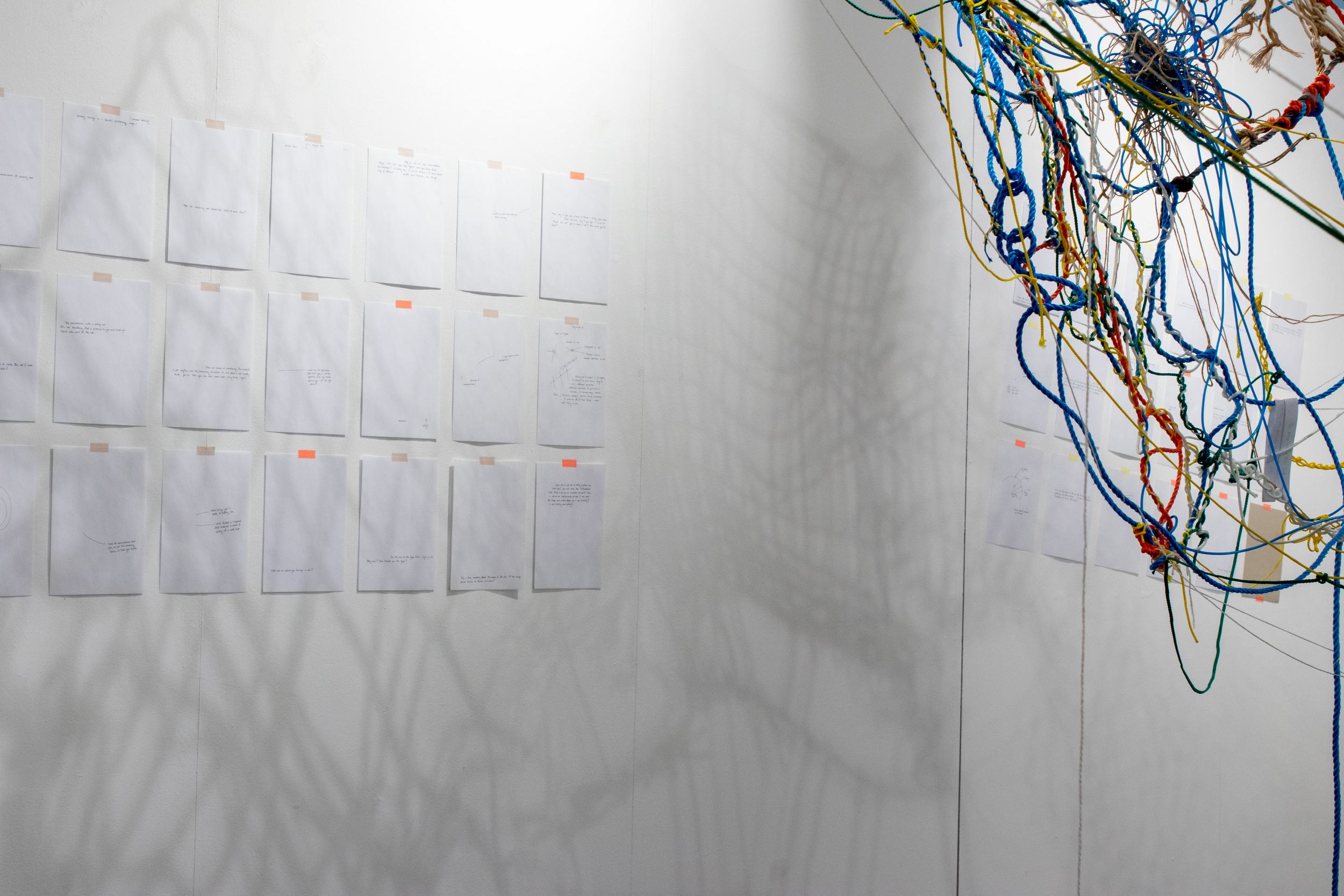 On the left, A4 papers are hanged to the wall with oranger and light pink tape. They are hanged in 3 rows to form a large grid, 6 and a half sheets are visible here. The papers are printed with sentences in handwriting and sketches. On the right, the corner of a net made of brightly colored ropes is visible. Behind the net hands another grid of papers, the grid is not horizontal but instead leaning down to the right.