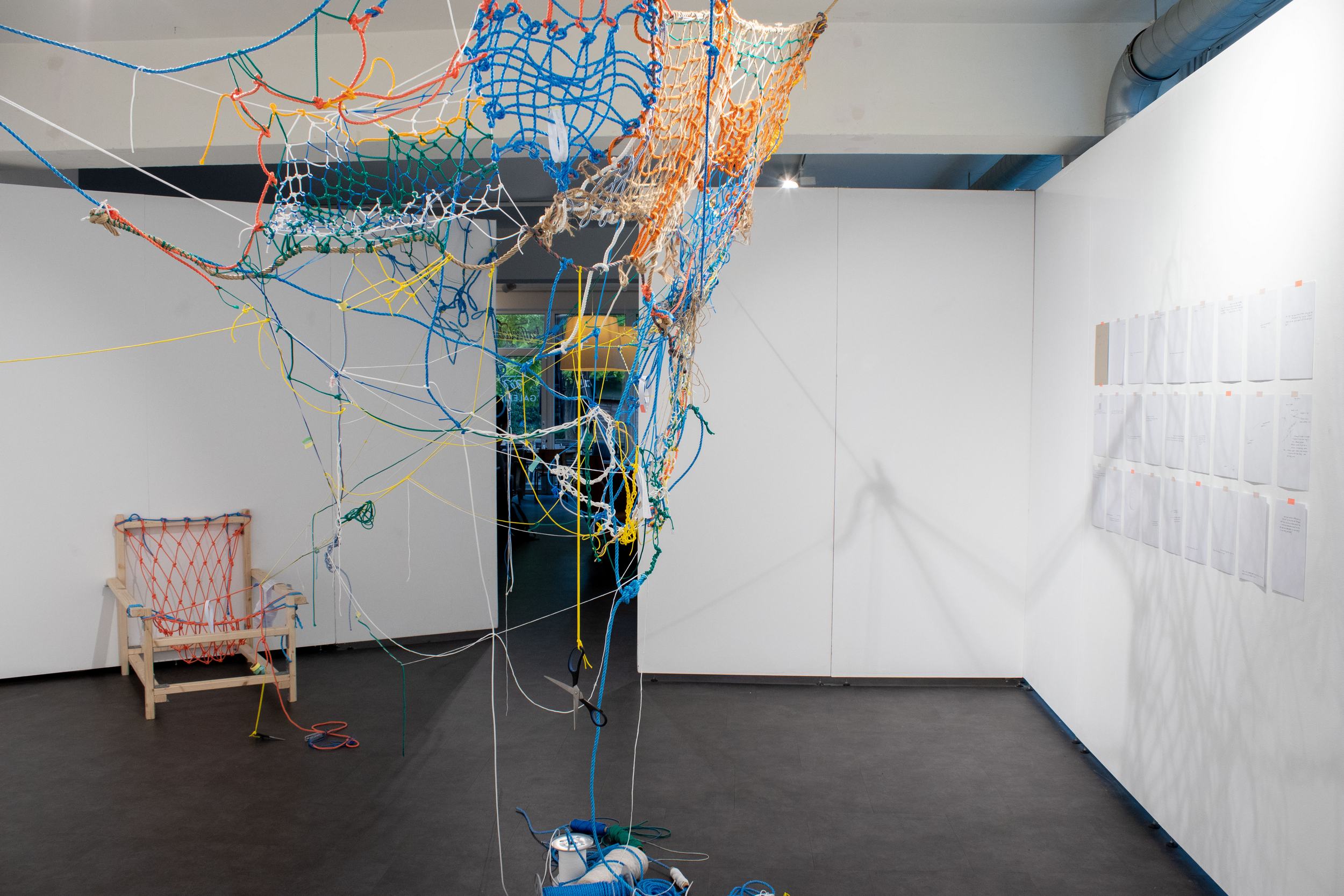 At the center, a large net made of bright colored ropes, blue and orange are dominant. On the right, a grid of papers hanged to the wall on 3 lines of 8 papers, forming a grid. On the left, behind the net, there is an armchair made of a bright orange net hanged on a wooden frame. Visitors have connected it to the net by a thin rope.