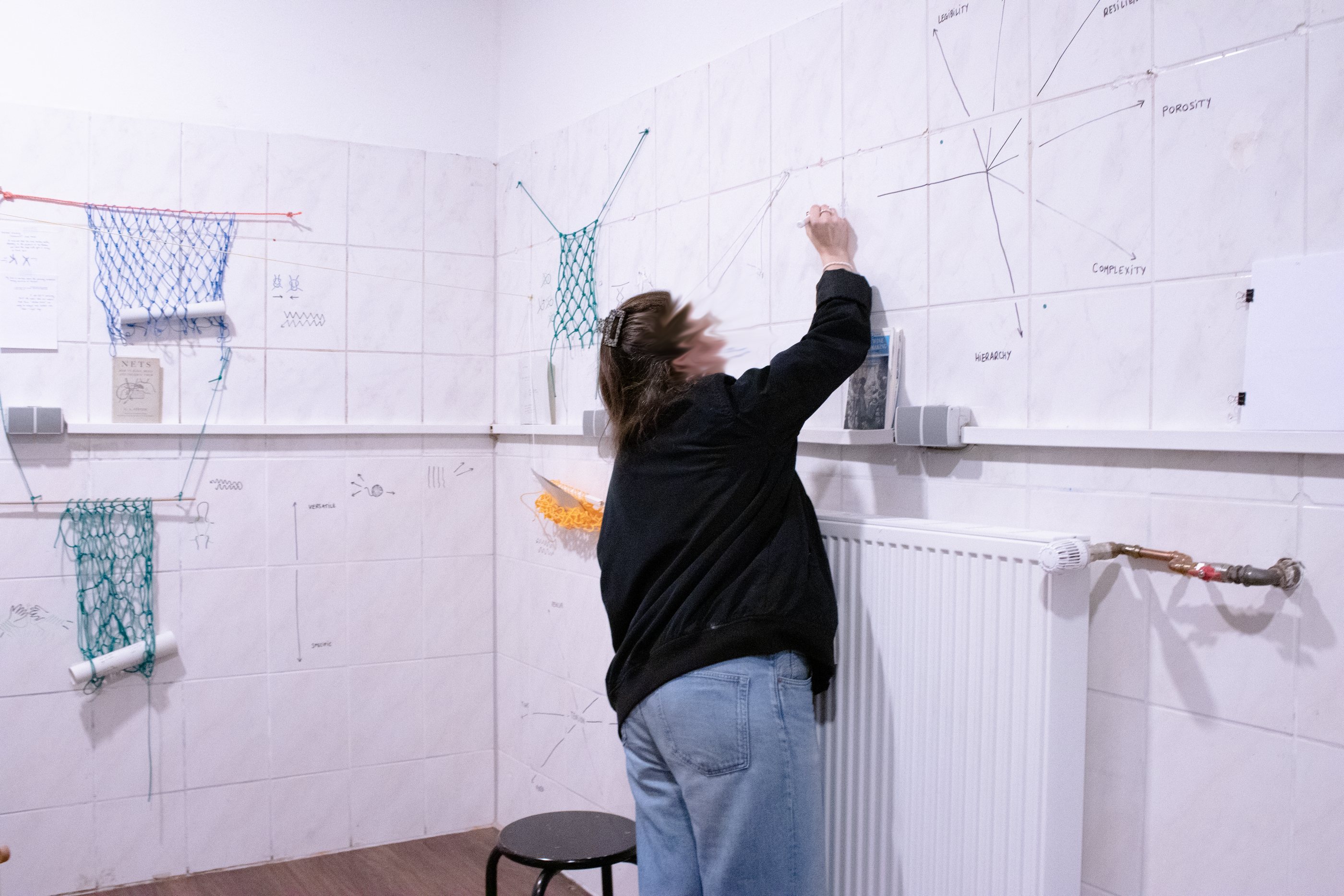 A person is writing on a tiled wall. In front of them there is a sketch with arrows starting at the center that point to words written in a circle, like 